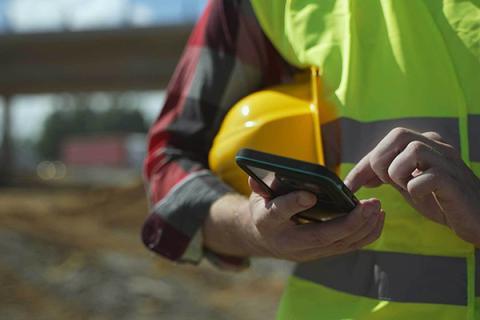 A worker in a yellow safety vest holds a hard hat under their arm while typing on a cell phone